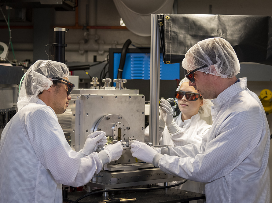 Three researchers in clean room attire adjust a Cell Cartridge Inserter at the end of the BELLA PW Proton beam line.