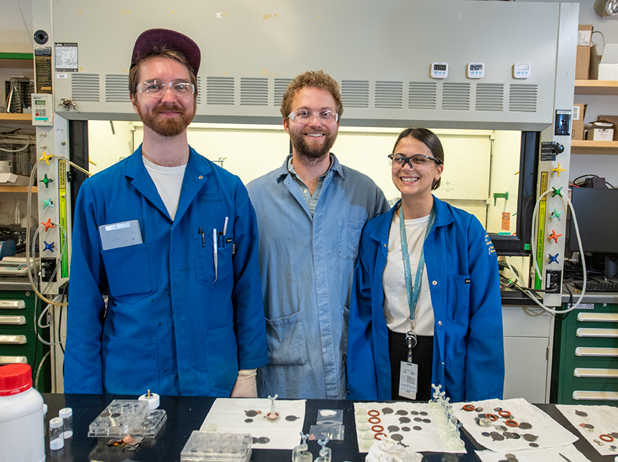 Three people in lab coats stand side by side, smiling in a laboratory setting.