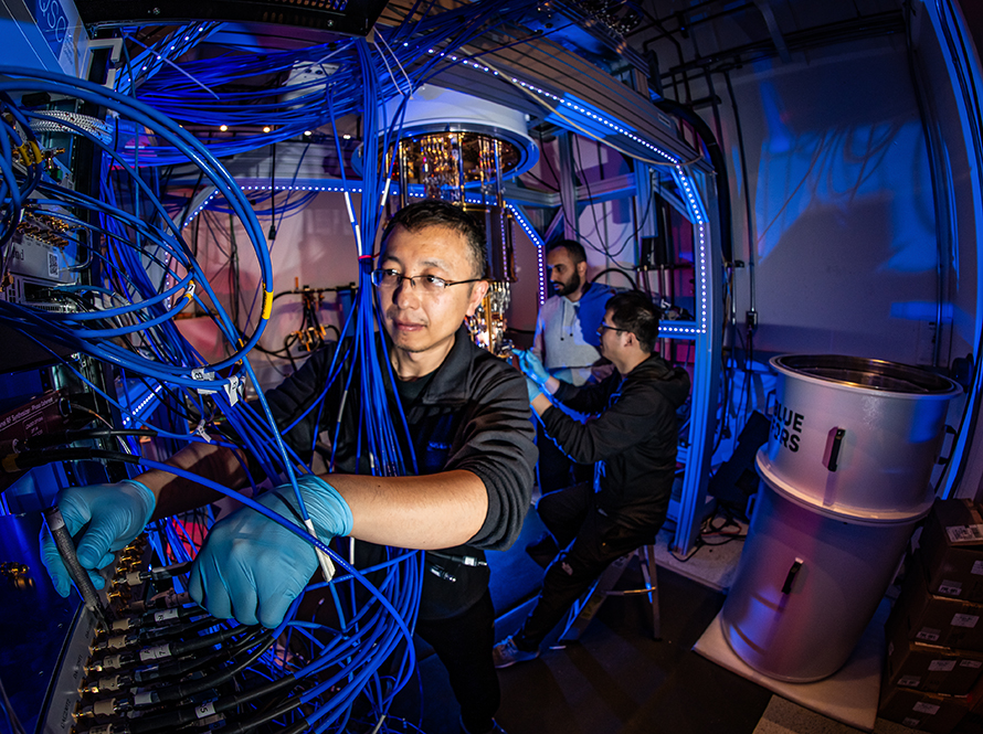 Gang Huang adjusts a quantum computer control unit while Ahmed Hajr and Ke Wang work on a dilution refrigerator in a lab.