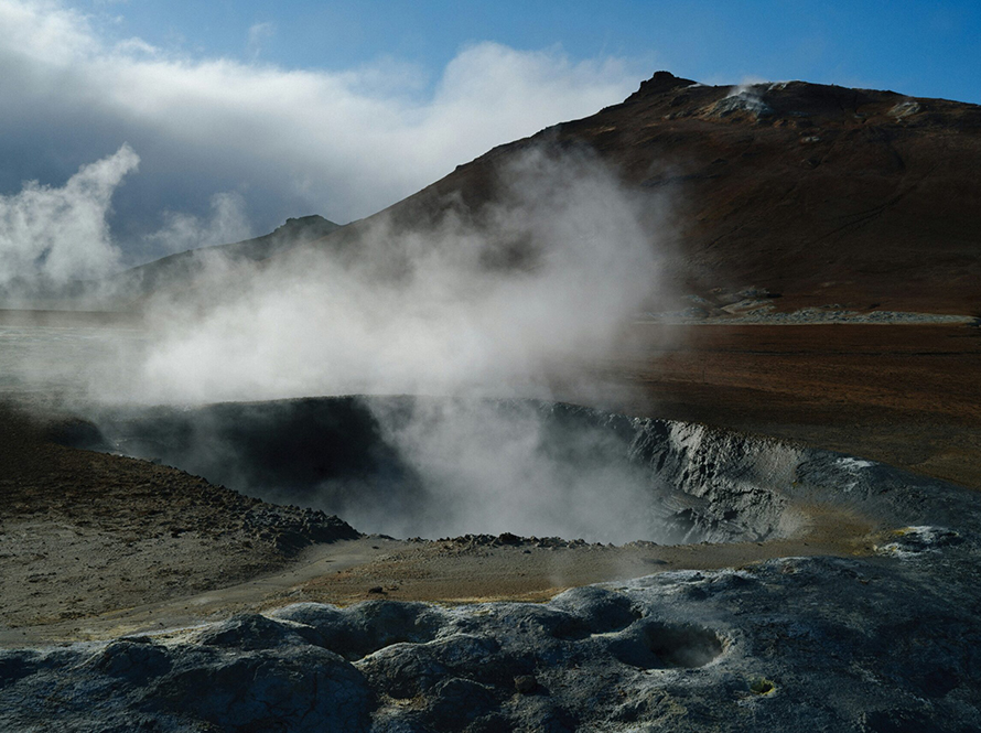 A large steam vent releasing white steam into the air, surrounded by rocky terrain.